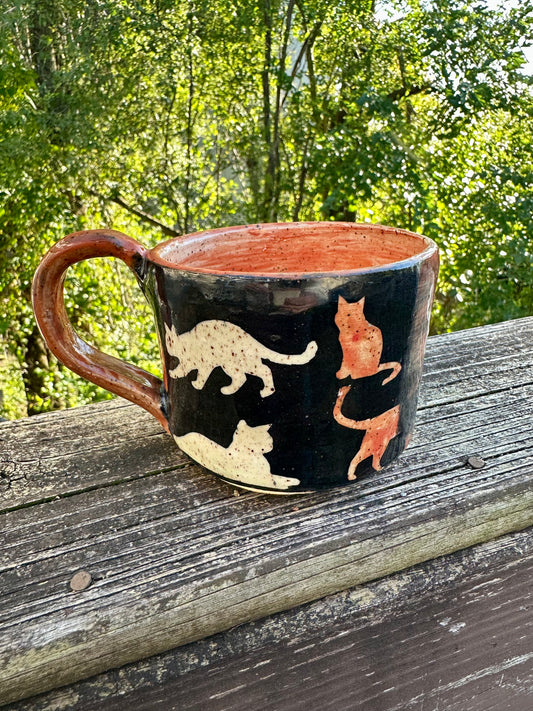 A handmade ceramic mug with a black exterior decorated with cat silhouettes in cream and orange, featuring an orange handle and interior, set on a weathered wooden railing with a lush green forest background.