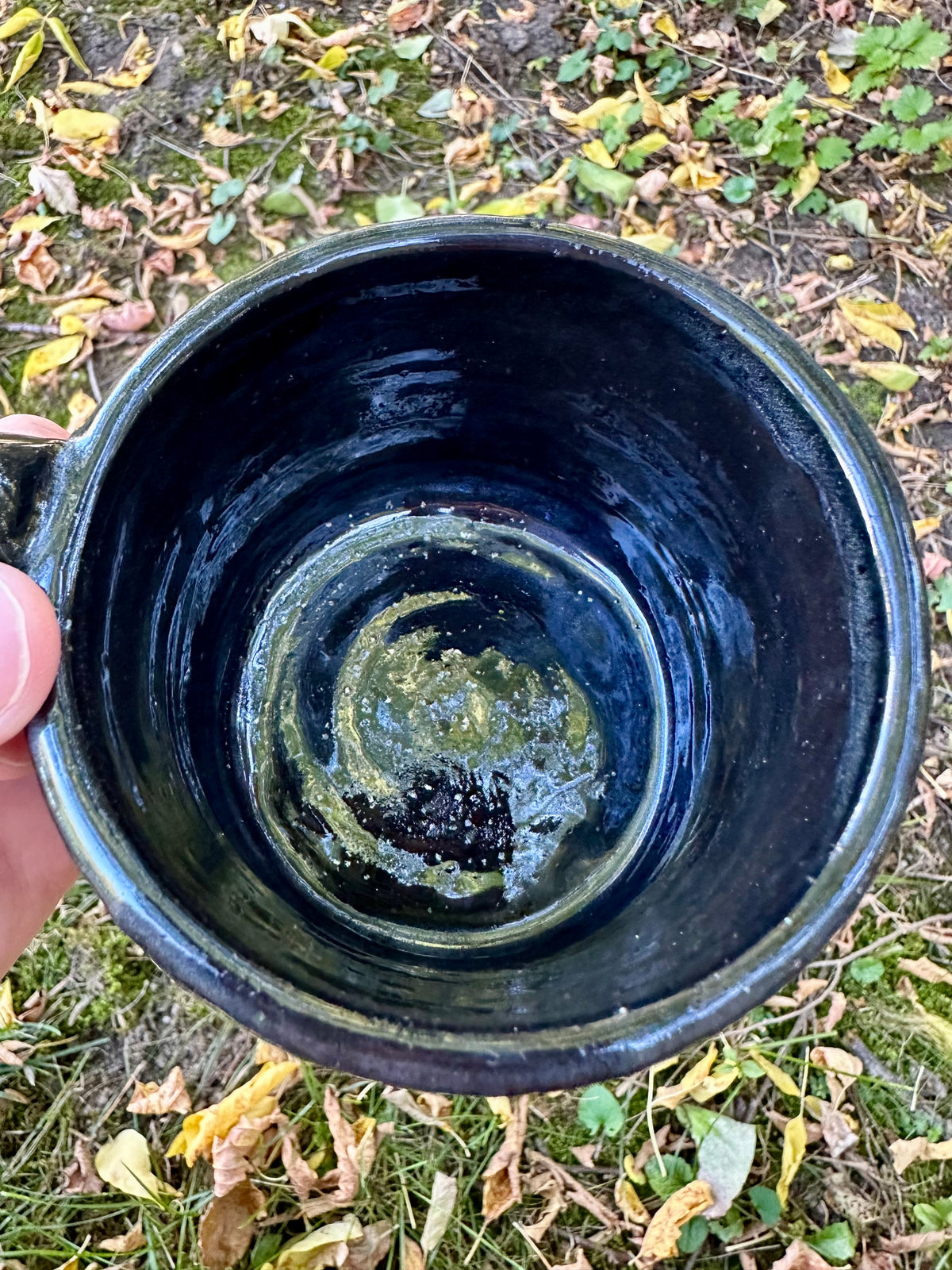 Interior shot of a handmade glossy black glazed ceramic mug with a subtle sparkle, held by a hand against a background of grass and fallen autumn leaves.