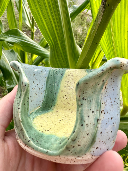 A hand holds a utensil holder themed as a corn stalk against a sky blue base; set in a garden in front of corn stalks.