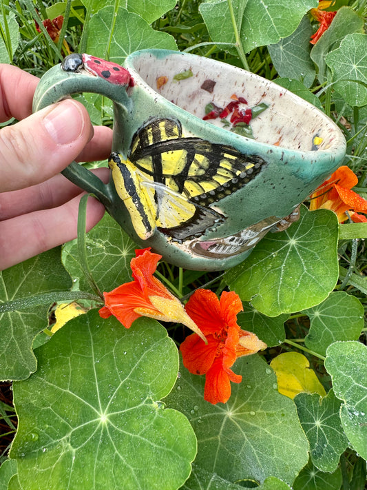 A hand holds a handmade green ceramic mug decorated with a yellow swallowtail butterfly and a partial brown Polyphemus moth and a sculpted ladybug on the top of the handle; set against a garden background with orange nasturtiums.