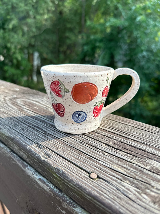 A speckled beige ceramic tea cup is decorated with painted red cherries, blueberries, strawberries and peaches, sitting on a worn wood rail set against a blurred green outdoor background.