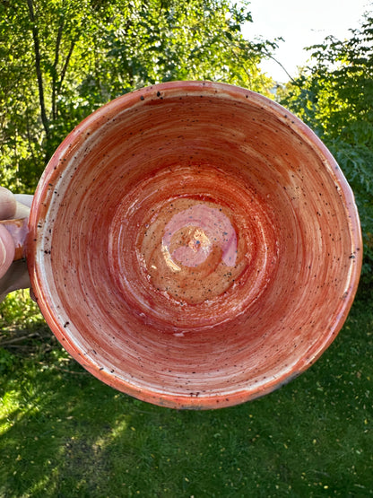 A handmade ceramic mug with a handle, shown from a top-down perspective held by a hand against a background of green grass and trees. The mug features a warm terracotta orange and reddish-brown glaze with subtle speckles.