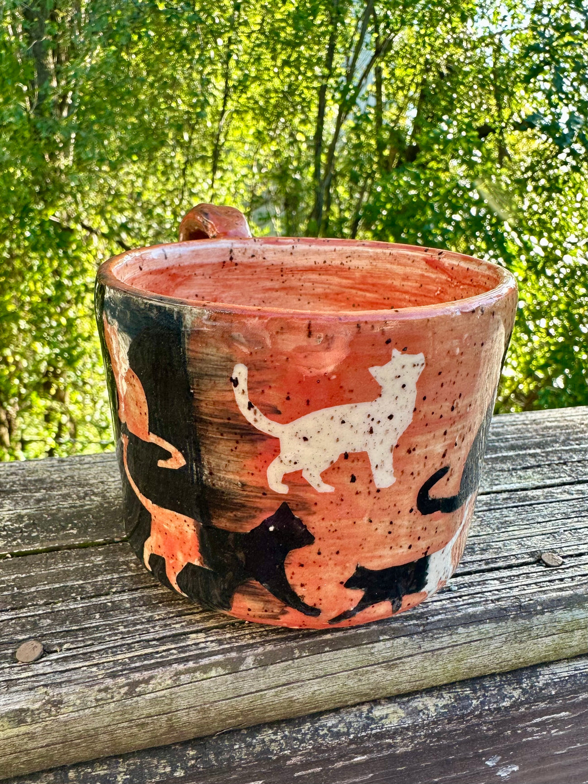 A handmade ceramic mug with am exterior transitioning from black to orange, decorated with cat silhouettes in black, cream and orange, featuring an orange handle and interior, set on a weathered wooden railing with a lush green forest background.