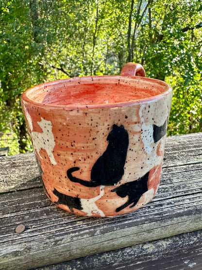 A handmade ceramic mug with a mostly orange exterior transitioning to white decorated with cat silhouettes in cream, black and orange, featuring an orange handle and interior, set on a weathered wooden railing with a lush green forest background.