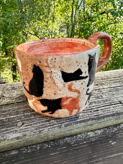A handmade ceramic mug with a mostly orange exterior transitioning to white decorated with cat silhouettes in cream, black and orange, featuring an orange handle and interior, set on a weathered wooden railing with a lush green forest background.