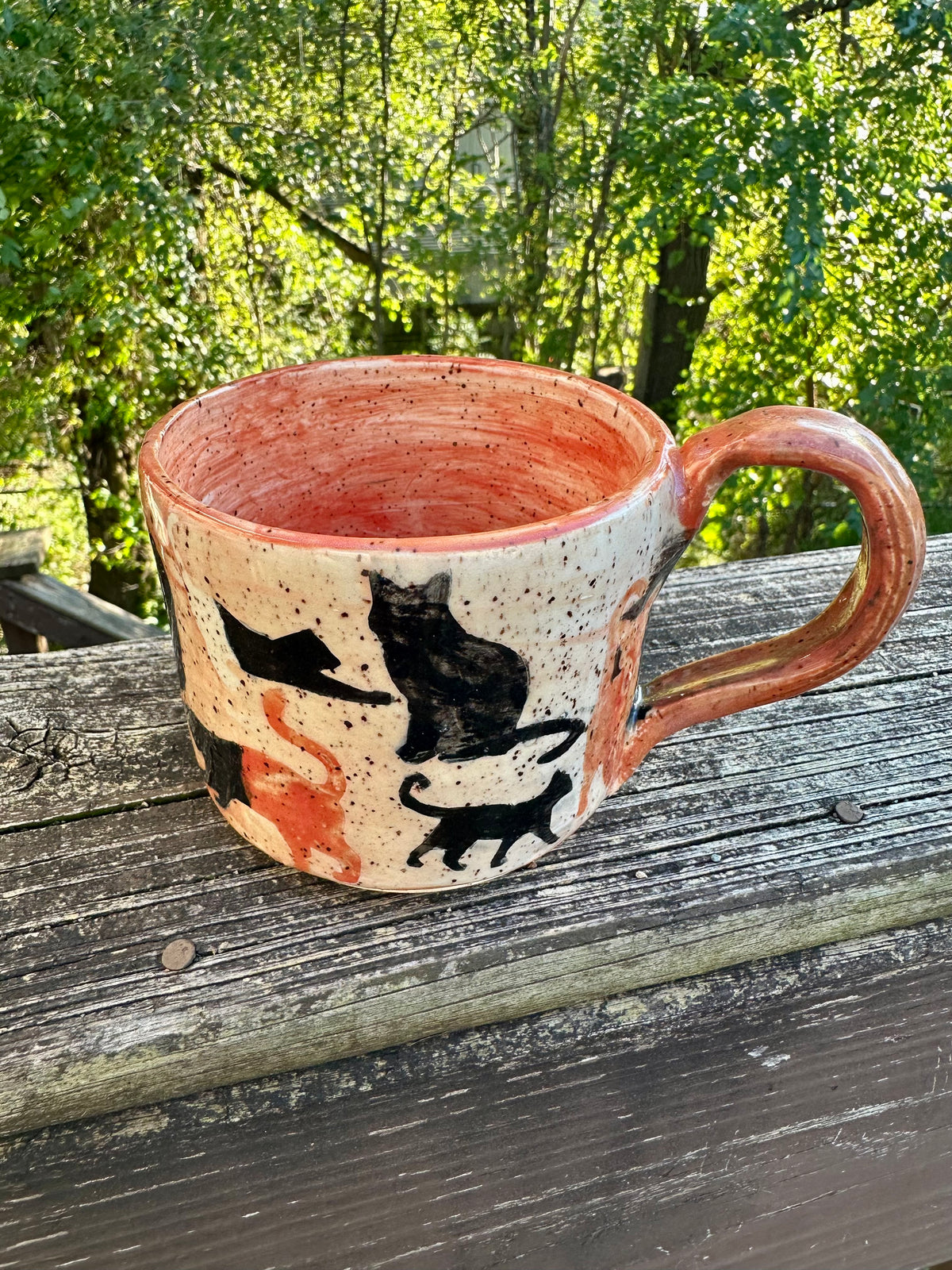 A handmade ceramic mug with an exterior transitioning from orange to white to black, decorated with cat silhouettes in black, cream and orange, featuring an orange handle and interior, set on a weathered wooden railing with a lush green forest background.