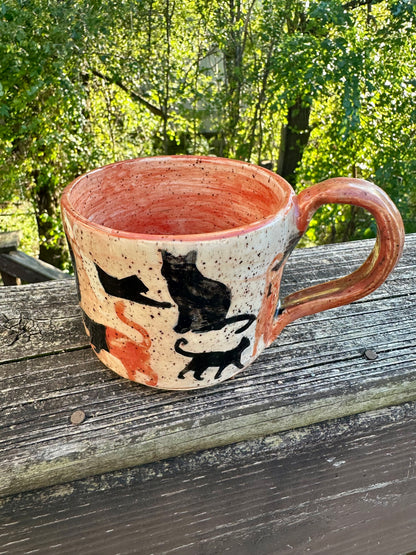 A handmade ceramic mug with an exterior transitioning from orange to white to black, decorated with cat silhouettes in black, cream and orange, featuring an orange handle and interior, set on a weathered wooden railing with a lush green forest background.