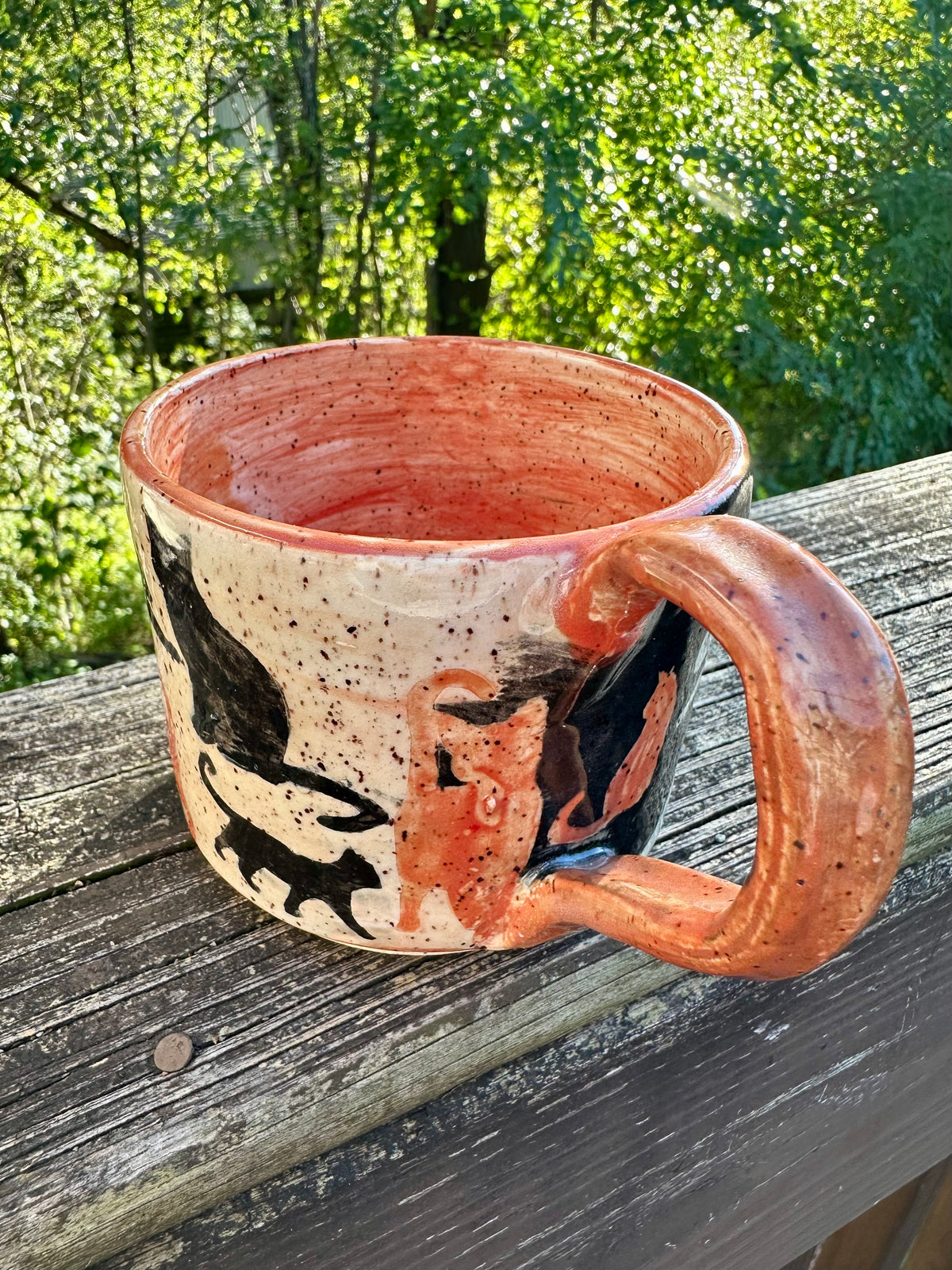 A handmade ceramic mug with an exterior transitioning from white to black, decorated with cat silhouettes in black and orange, featuring an orange handle and interior, set on a weathered wooden railing with a lush green forest background.
