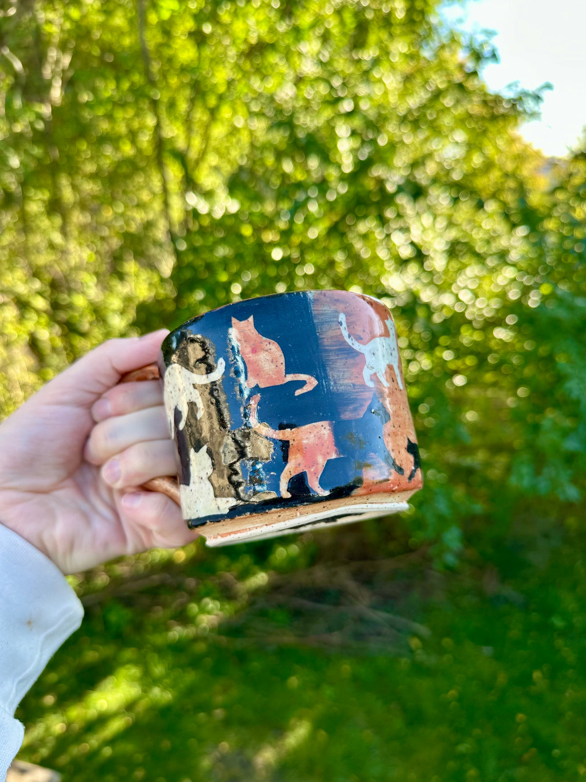 A hand holding a handmade ceramic mug with an exterior transitioning from black to orange with orange, white, and black painted cat illustrations; set against a blurred green outdoor background.