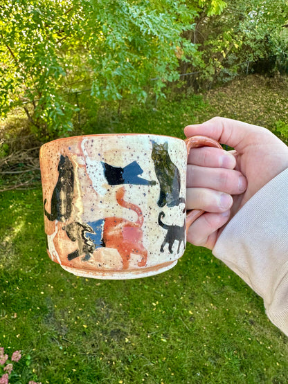 A hand holding a handmade ceramic mug outdoors in a grassy garden with fall leaves in the background. The mug has an orange fading to cream exterior with painted cat silhouettes in black, orange, and white.