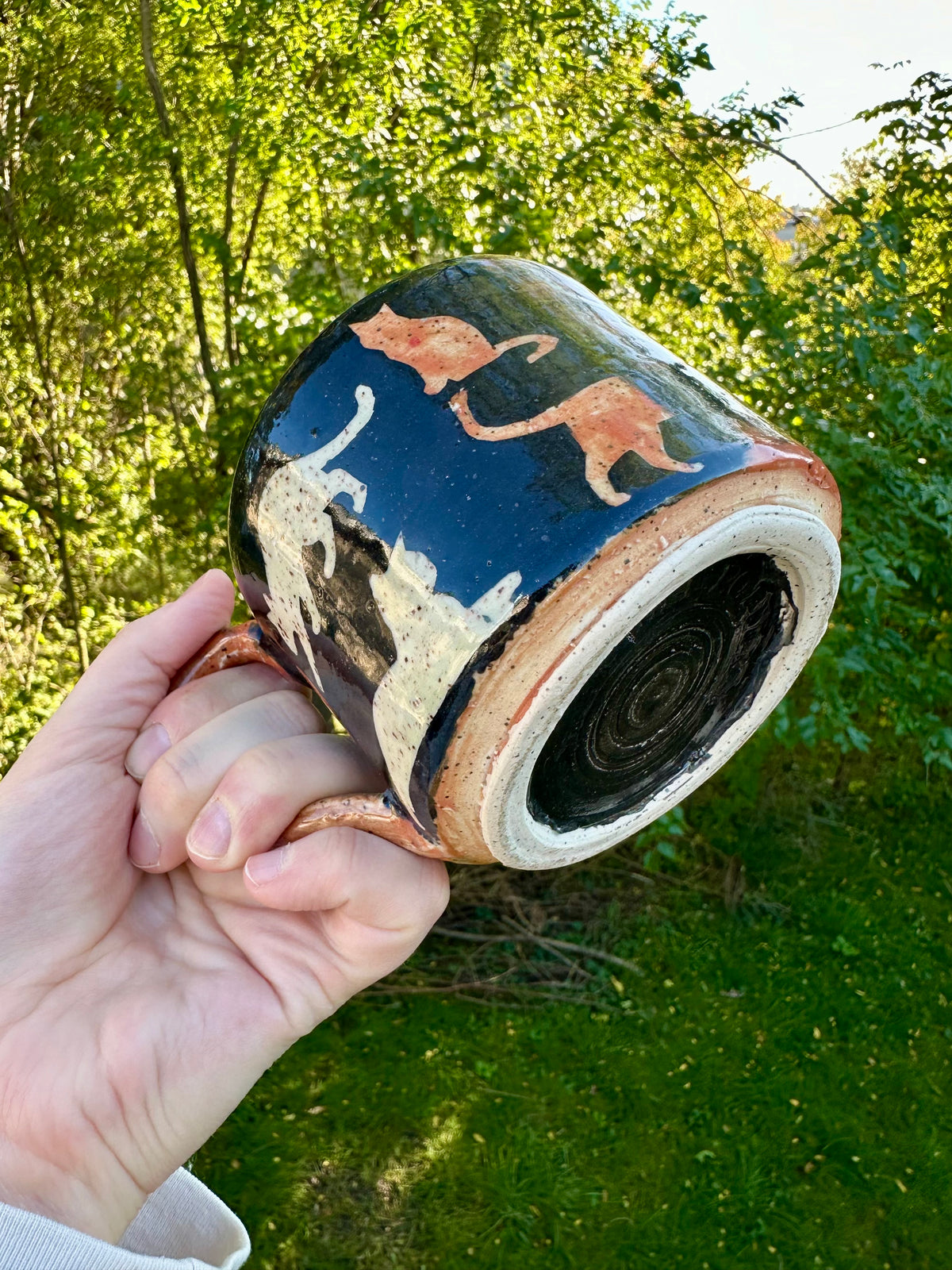 A hand holding a handmade ceramic mug with a black exterior with orange, white, and black painted cat illustrations and an orange, white, and black bottom; set against a blurred green outdoor background.