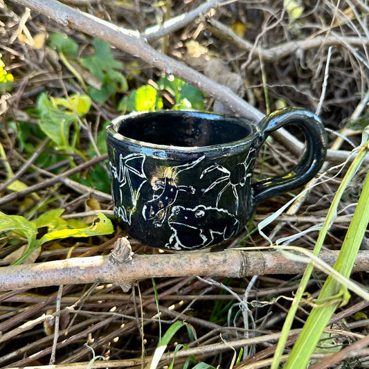 A small black ceramic cup with white line-art illustrations of figures and animals, placed outdoors on a branch surrounded by dry twigs and green leaves.