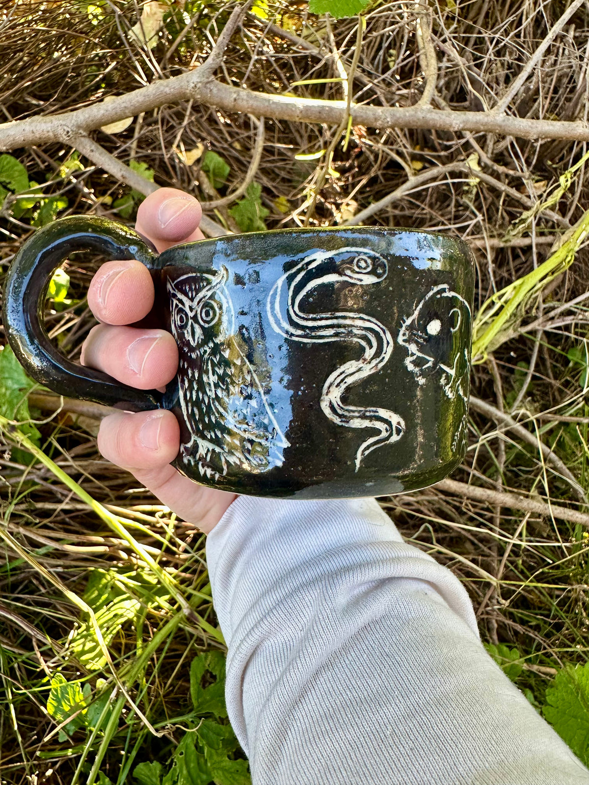 A small black ceramic cup with white line-art illustrations of figures and animals, placed outdoors on a branch surrounded by dry twigs and green leaves.