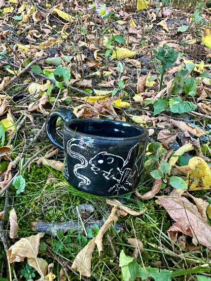 A small black ceramic cup with white line-art illustrations of figures and animals, placed outdoors on a branch surrounded by dry twigs and green leaves.