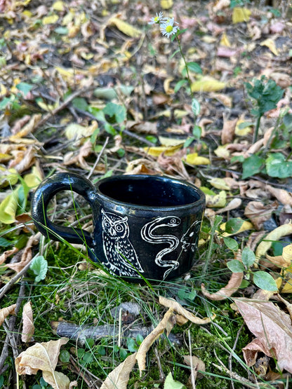 A small black ceramic cup with white line-art illustrations of figures and animals, placed outdoors on a branch surrounded by dry twigs and green leaves.