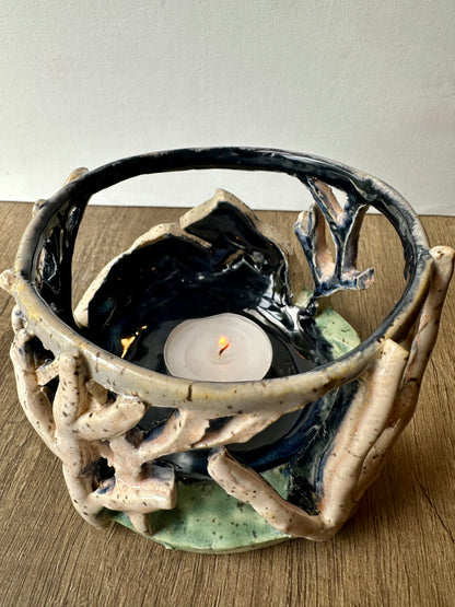 Round ceramic candle surround with mountain and birch trees cut out; lit tea light candle placed inside.
