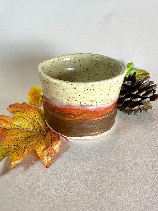 A handmade ceramic cup with a speckled cream top, a red-orange band, and a brown base, styled with autumn leaves, a pinecone, and a small yellow flower on a neutral background.