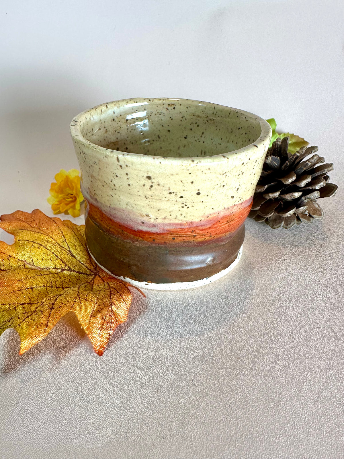 A handmade ceramic cup with a speckled cream top, a red-orange band, and a brown base, styled with autumn leaves, a pinecone, and a small yellow flower on a neutral background.