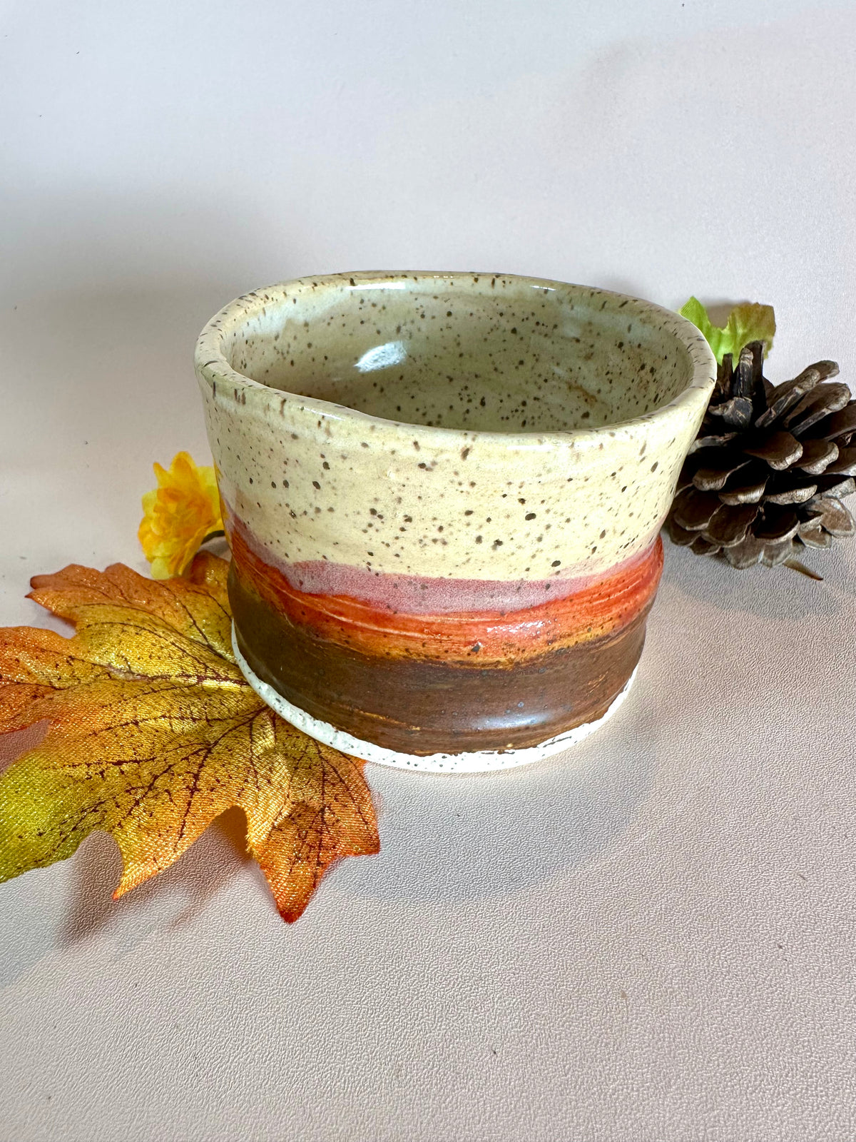 A handmade ceramic cup with a speckled cream top, a red-orange band, and a brown base, styled with autumn leaves, a pinecone, and a small yellow flower on a neutral background.