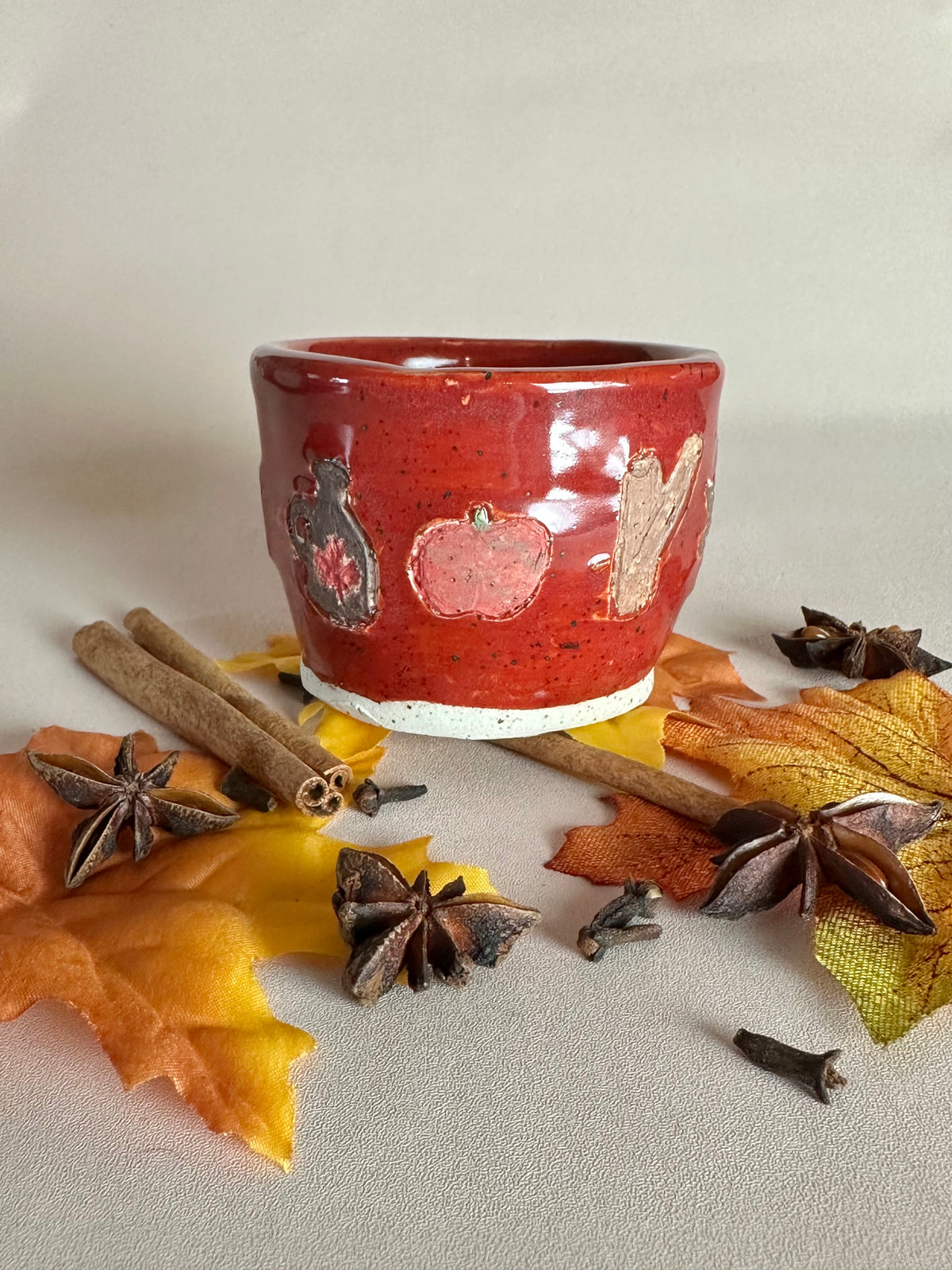 A small handmade ceramic cup with a glossy red glaze and hand-painted apples, maple syrup jug, and cinnamon; styled with cinnamon sticks, cloves, and fall leaves on a neutral background.