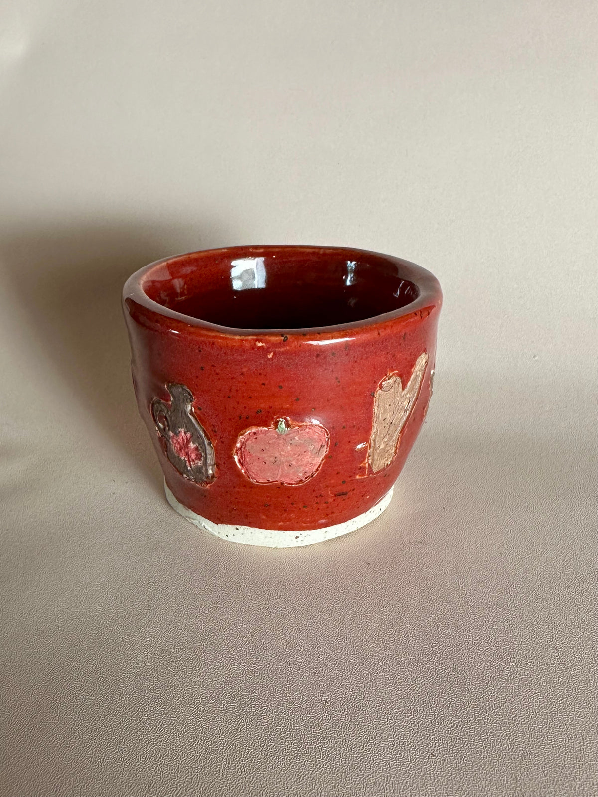 A small handmade ceramic cup with a glossy red glaze and hand-painted apples, maple syrup jug, and cinnamon; styled on a neutral background.