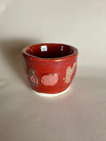 A small handmade ceramic cup with a glossy red glaze and hand-painted apples, maple syrup jug, and cinnamon; styled on a neutral background.