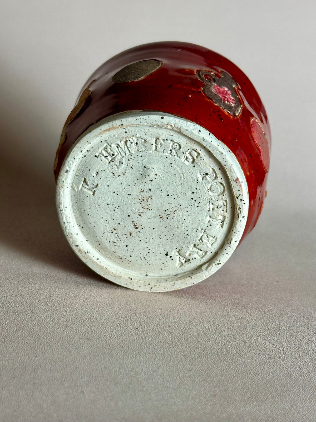 A small handmade ceramic cup with a glossy red glaze and hand-painted apples, maple syrup jug, and whole nutmeg; tilted on its side, showing the black-speckled white clay bottom with "K. Embers Pottery" stamped; styled on a neutral background. 