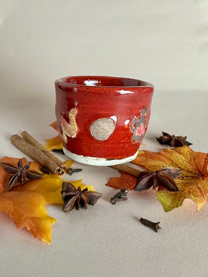 A small handmade ceramic cup with a glossy red glaze and hand-painted orange peel, whole nutmeg, and maple syrup jug; styled with cinnamon sticks, cloves, and fall leaves on a neutral background.