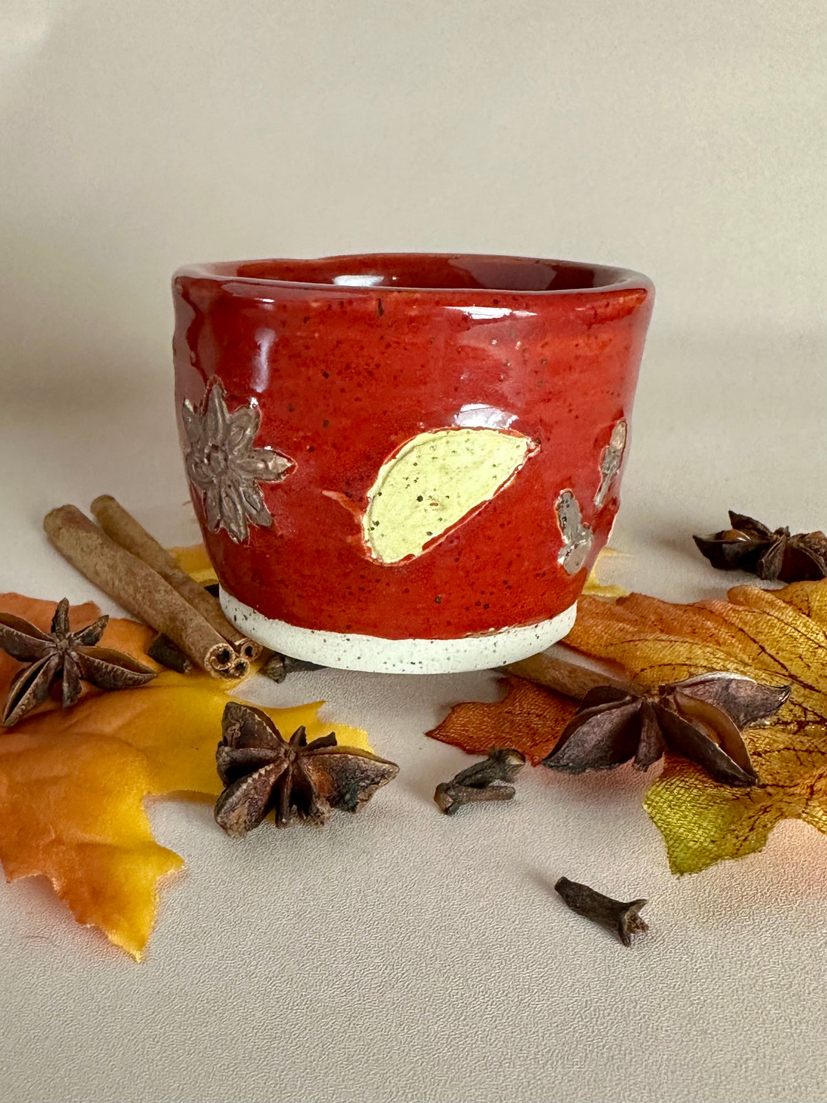 A small handmade ceramic cup with a glossy red glaze and hand-painted apples slice, star anise, and cloves; styled with cinnamon sticks, cloves, and fall leaves on a neutral background.