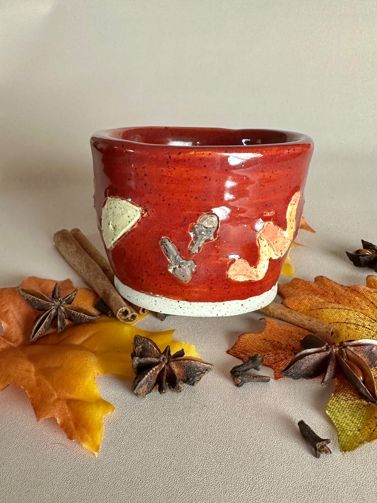 A small handmade ceramic cup with a glossy red glaze and hand-painted apple slice, cloves, and orange peel; styled with cinnamon sticks, cloves, and fall leaves on a neutral background.