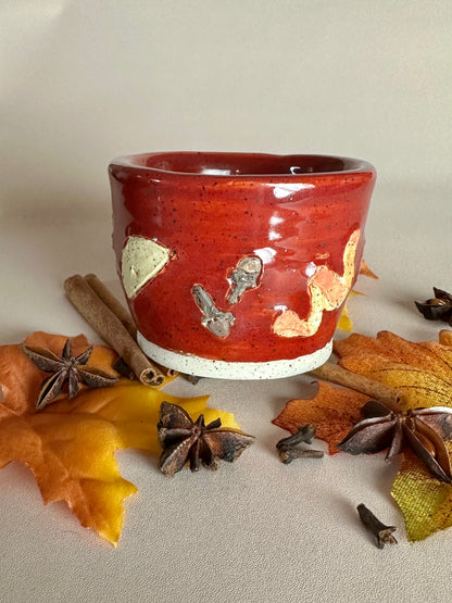 A small handmade ceramic cup with a glossy red glaze and hand-painted apple slice, cloves, and orange peel; styled with cinnamon sticks, cloves, and fall leaves on a neutral background.