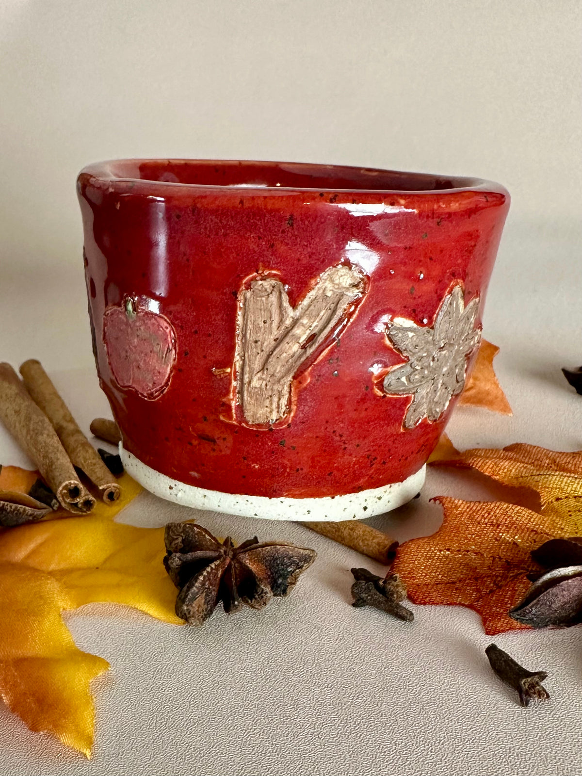 A small handmade ceramic cup with a glossy red glaze and hand-painted apples, cinnamon, and star anise; styled with cinnamon sticks, cloves, and fall leaves on a neutral background.