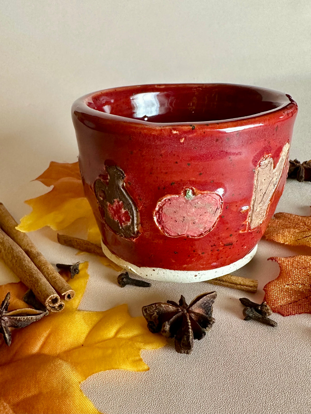 A small handmade ceramic cup with a glossy red glaze and hand-painted apples, maple syrup jug, and cinnamon; styled with cinnamon sticks, cloves, and fall leaves on a neutral background.