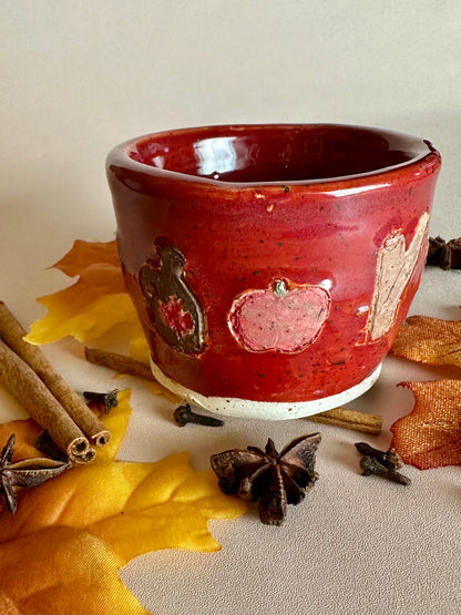 A small handmade ceramic cup with a glossy red glaze and hand-painted apples, maple syrup jug, and cinnamon; styled with cinnamon sticks, cloves, and fall leaves on a neutral background.