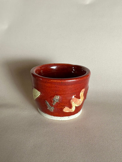 A small handmade ceramic cup with a glossy red glaze and hand-painted apple slice, cloves, and orange peel; styled on a neutral background.