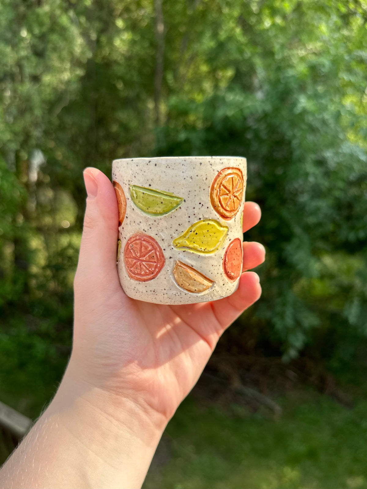 A hand holds a small speckled white ceramic cup with colorful whole citrus fruits, citrus wedges, and citrus slices illustrations in front of a blurred green garden background.