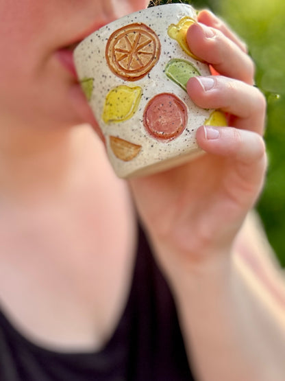 A close-up lifestyle shot of a person holding a small speckled ceramic cup decorated with colorful embossed citrus slices, with a blurred green outdoor background.