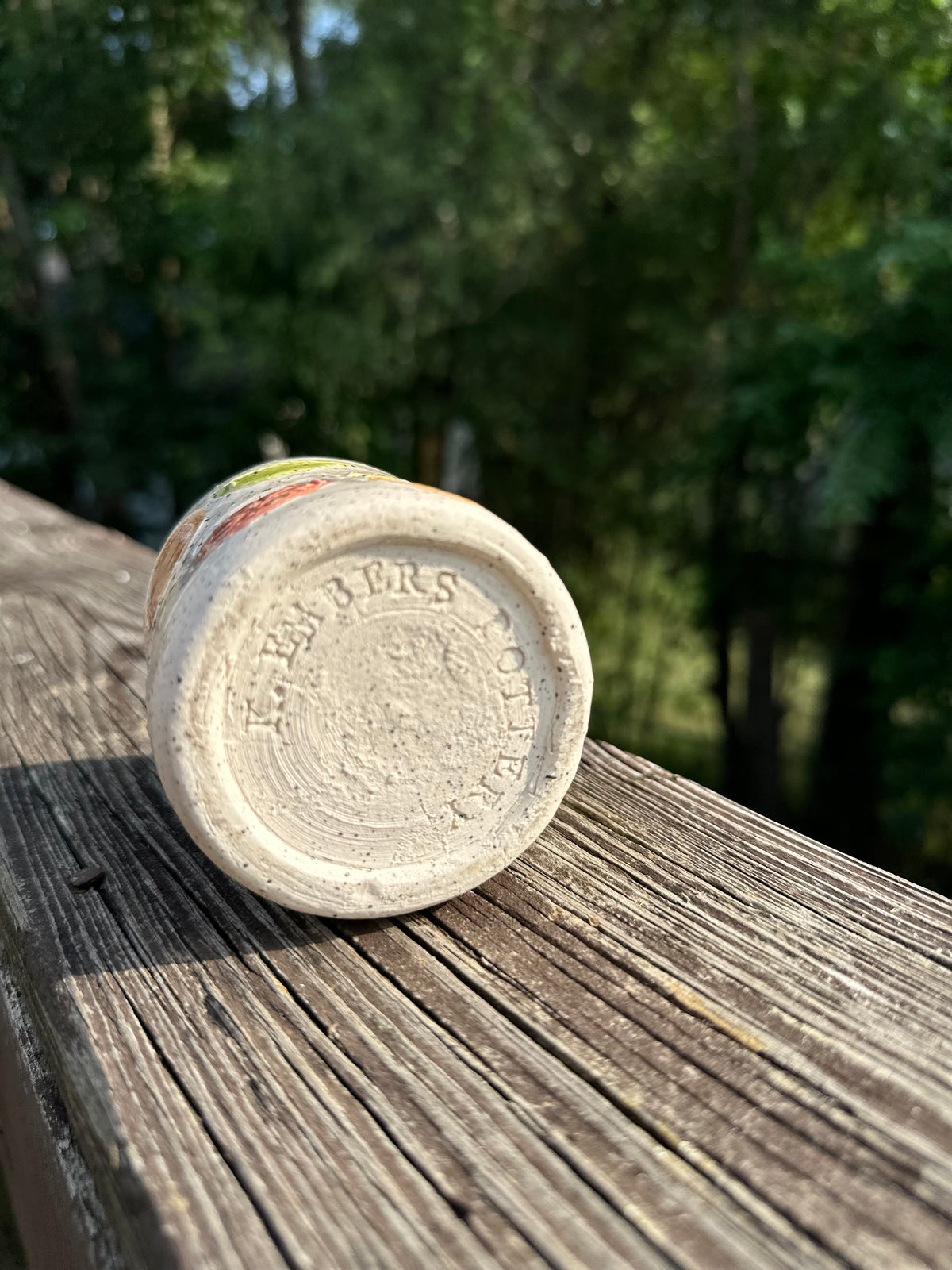 The bottom of a speckled white ceramic cup with "K. Embers Pottery" stamped into the base, on a worn wooden deck rail in front of a blurred green forest background.