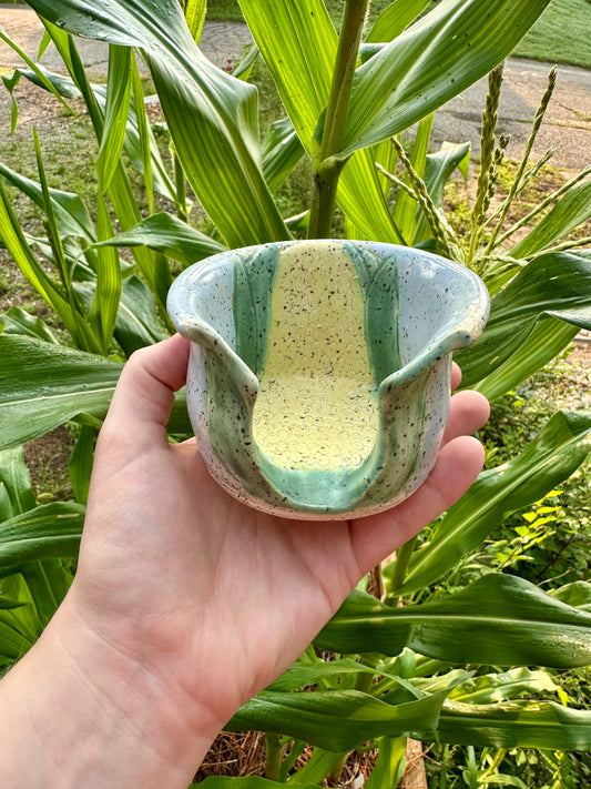 A hand holds a utensil holder themed as a corn stalk against a sky blue base; set in a garden in front of corn stalks.