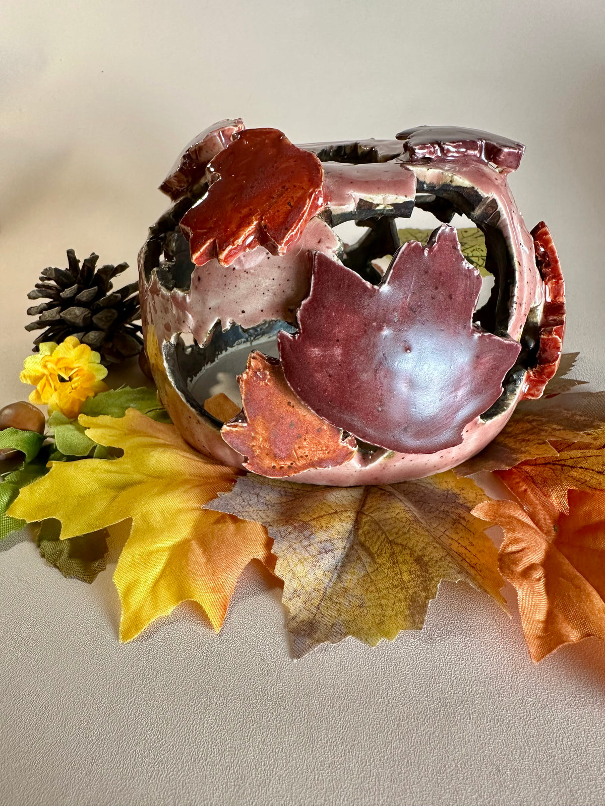 A decorative ceramic sphere with glossy mauve base glaze, featuring maple leaf-shaped cutouts in various shades of red, styled on artificial autumn leaves and a pinecone against a neutral beige background.