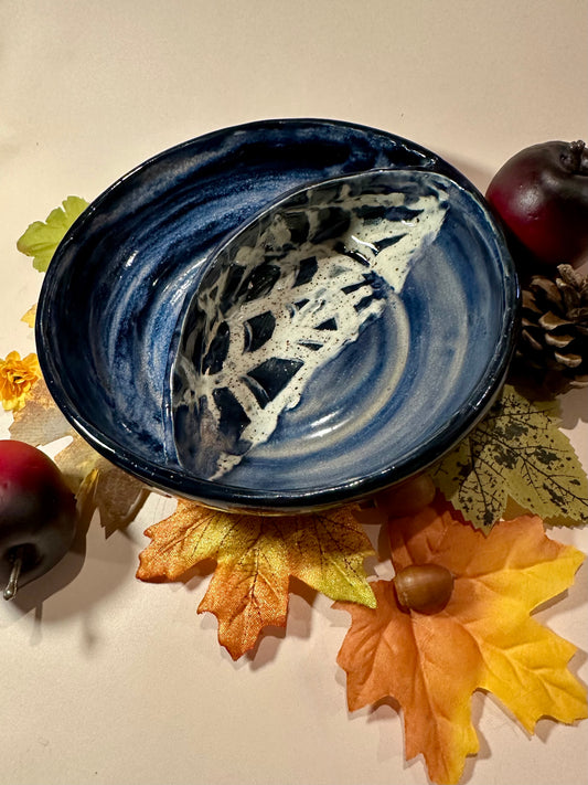 A ceramic serving bowl with a deep blue and white marbled swirls with half-spider web pattern, styled with autumn leaves, acorns, and red apples on a beige background.