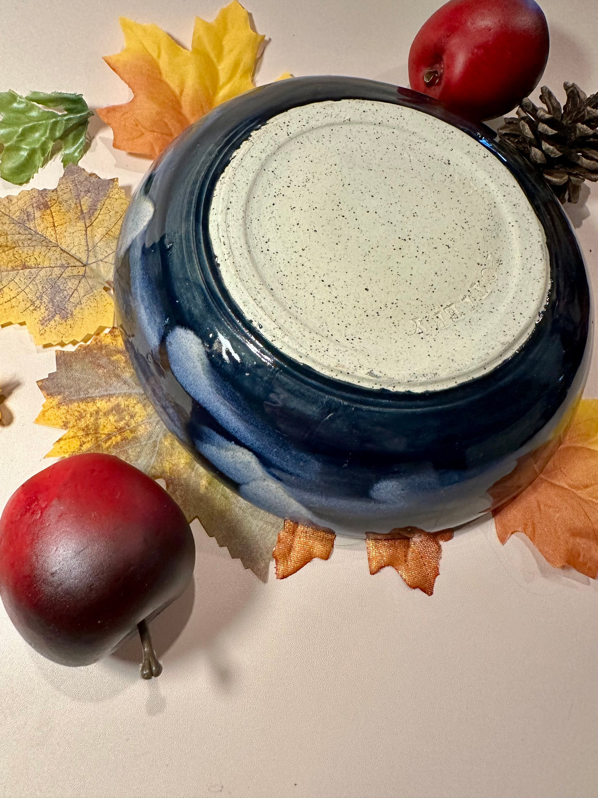 A round ceramic bowl with a glossy deep blue exterior and a speckled off-white bottom; styled with autumn leaves, pinecones, and red apples on a light surface.