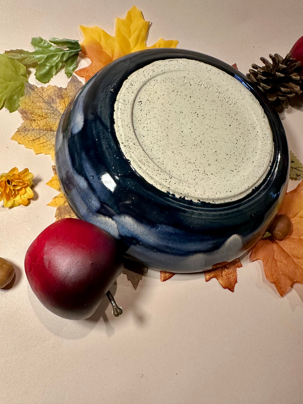A round ceramic bowl with a glossy deep blue exterior and a speckled off-white bottom; styled with autumn leaves, pinecones, and red apples on a light surface.