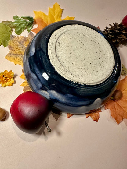 A round ceramic bowl with a glossy deep blue exterior and a speckled off-white bottom; styled with autumn leaves, pinecones, and red apples on a light surface.