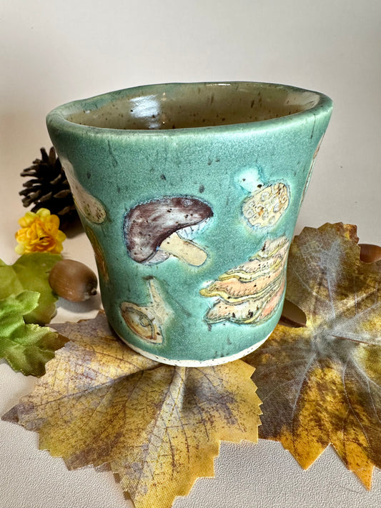 A jade green ceramic cup with brown, orange, yellow, tan, and white mushroom illustrations, stylized with fall foliage underneath on a solid neutral backdrop.
