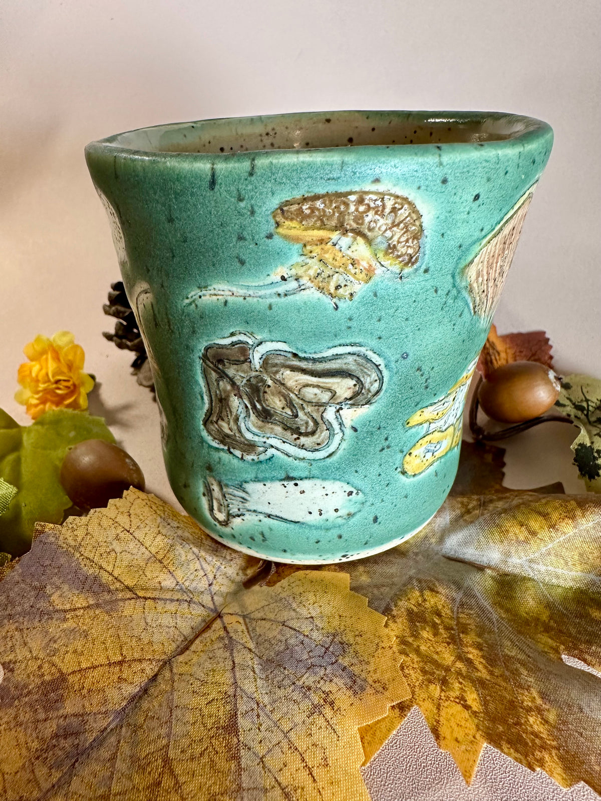 A jade green ceramic cup with brown, orange, yellow, tan, and white mushroom illustrations, stylized with fall foliage underneath on a solid neutral backdrop.