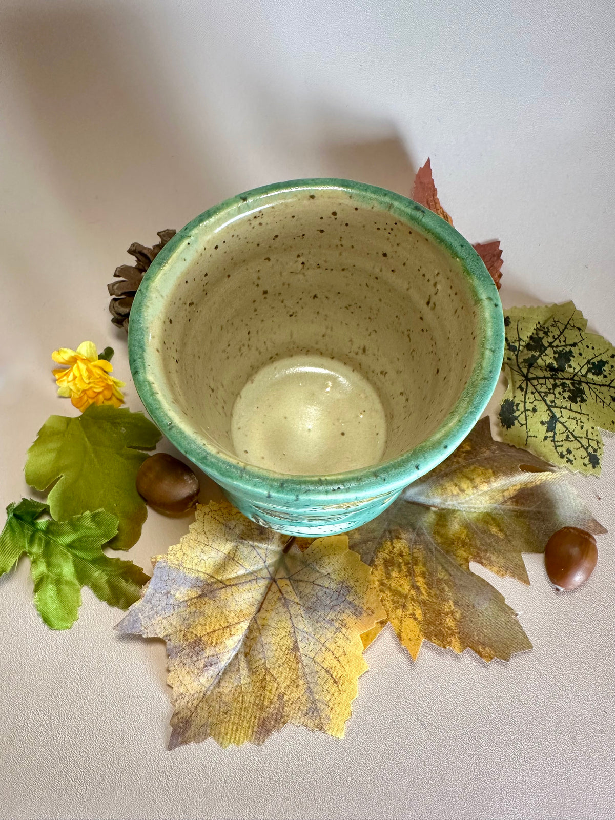 Interior of a jade green ceramic cup showing a glossy speckled tan interior, stylized with fall foliage underneath on a solid neutral backdrop.