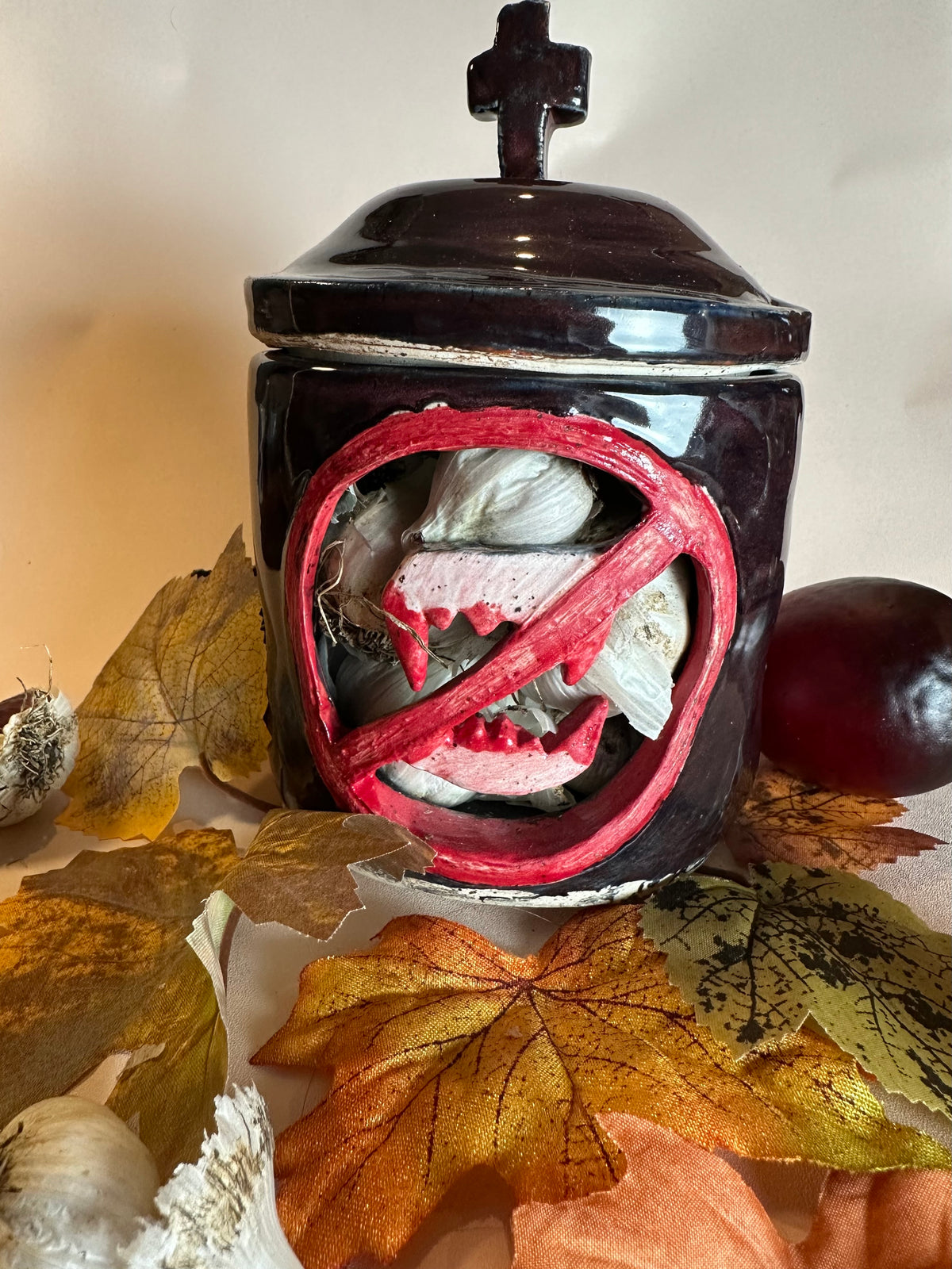 A glossy black canister filled with garlic, a cut out of a set of bloody vampire fangs inside a red circle with a slash; topped with a lid with a cross as a handle. Styled with fall leaves, an apple, and garlic on a solid neutral background.