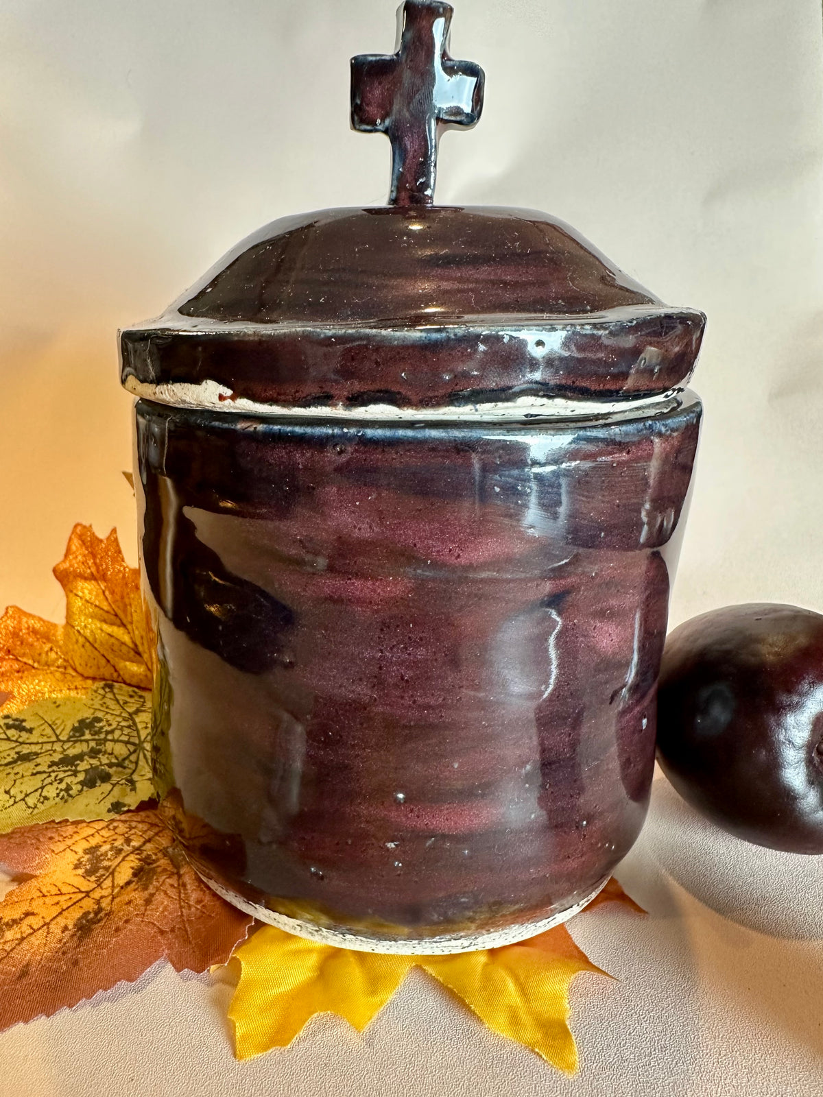 A glossy black canister topped with a lid with a cross as a handle. Styled with fall leaves and an apple on a solid neutral background.
