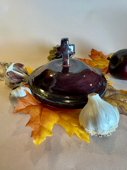 A glossy black canister lid with a cross as a handle. Styled with fall leaves, an apple, and garlic on a solid neutral background.